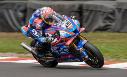Motorcycle racer in blue, red, and white livery leaning into a turn on a race track, helmeted rider in full gear behind a blue race bike with number plate visible on the front fairing, red and white curb in the foreground.