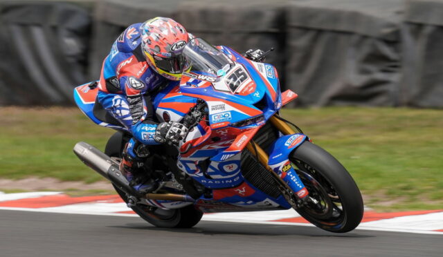 Motorcycle racer in blue, red, and white livery leaning into a turn on a race track, helmeted rider in full gear behind a blue race bike with number plate visible on the front fairing, red and white curb in the foreground.