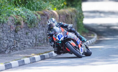 Motorcycle racer in full gear leaning into a sharp turn on a sunlit road beside a stone wall and greenery.