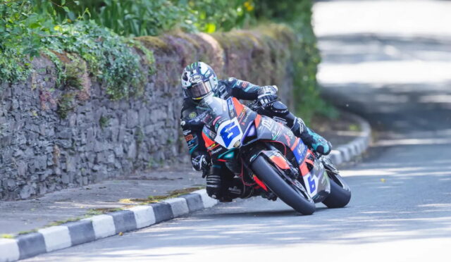 Motorcycle racer in full gear leaning into a sharp turn on a sunlit road beside a stone wall and greenery.