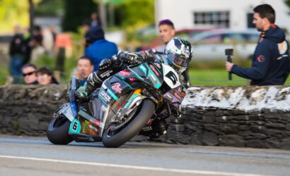 Rider in teal and black racing suit leans sharply into a turn on a road-race motorcycle, spectators and a stone barrier in the background.
