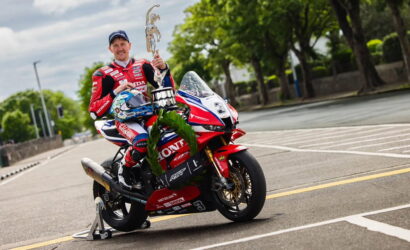 MotoGP rider in red Honda racing suit sits on a red Honda motorcycle, holding a trophy and leaping on an urban street post-race. laurel leaves hang on the bike.