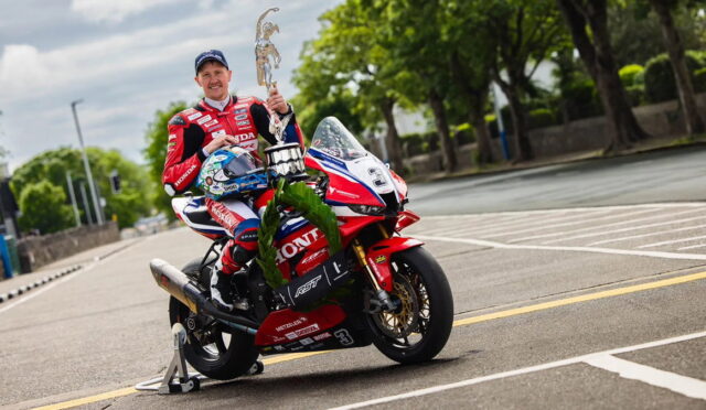 Racing rider in a red Honda suit sits on a red-and-white Honda motorcycle, holding a tall trophy on a street course with greenery in the background.