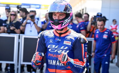 MotoGP rider in blue Pramac Yamaha suit, helmet on, giving thumbs up in the pit lane with photographers in the background nearby.