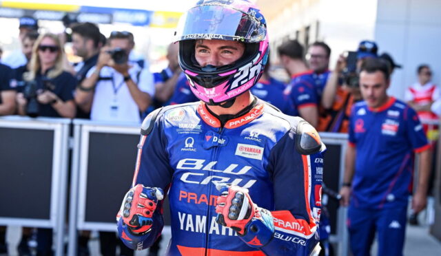 MotoGP rider in blue Pramac Yamaha suit, helmet on, giving thumbs up in the pit lane with photographers in the background nearby.
