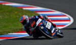 Two motorcycle racers leaning into a curve on a racetrack, bike number 56 in blue and orange fronting the pair.