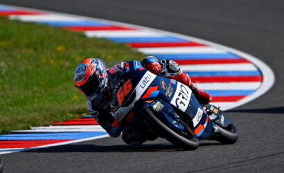 Two motorcycle racers leaning into a curve on a racetrack, bike number 56 in blue and orange fronting the pair.