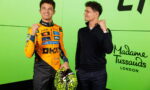 Two men posing together against a bright lime wall; the left man wears an orange racing suit with sponsor logos and holds a neon yellow helmet, while the right man in a black sweater makes a fist gesture and smiles.