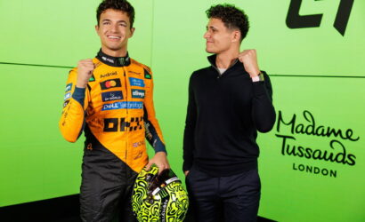 Two men posing together against a bright lime wall; the left man wears an orange racing suit with sponsor logos and holds a neon yellow helmet, while the right man in a black sweater makes a fist gesture and smiles.