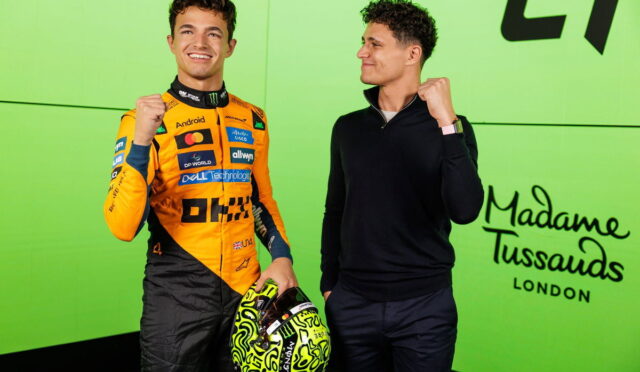 Two men posing together against a bright lime wall; the left man wears an orange racing suit with sponsor logos and holds a neon yellow helmet, while the right man in a black sweater makes a fist gesture and smiles.