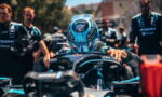 Race car driver in a turquoise helmet seated in the cockpit, hands on the steering wheel, surrounded by pit crew in teal uniforms.