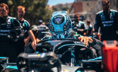 Race car driver in a turquoise helmet seated in the cockpit, hands on the steering wheel, surrounded by pit crew in teal uniforms.