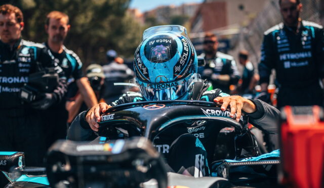 Race car driver in a turquoise helmet seated in the cockpit, hands on the steering wheel, surrounded by pit crew in teal uniforms.