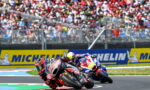 Two motorcycle racers lean into a tight turn on a race track as a crowded grandstand watches behind and Michelin banners line the wall.