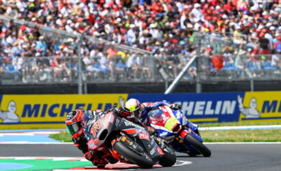 Two motorcycle racers lean into a tight turn on a race track as a crowded grandstand watches behind and Michelin banners line the wall.