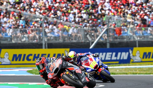 Two motorcycle racers lean into a tight turn on a race track as a crowded grandstand watches behind and Michelin banners line the wall.