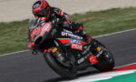 Racing motorcycle with a rider in red-black gear leaning into a turn on a track, sponsor logos visible on the bike.
