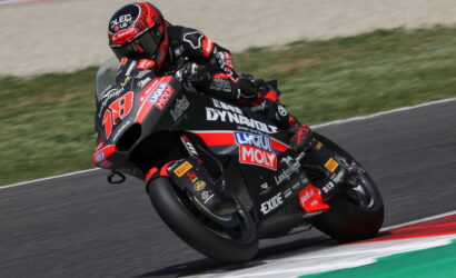 Racing motorcycle with a rider in red-black gear leaning into a turn on a track, sponsor logos visible on the bike.