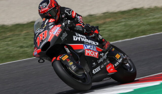 Racing motorcycle with a rider in red-black gear leaning into a turn on a track, sponsor logos visible on the bike.
