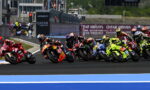 Group of motorcycle racers lean into a sharp turn on a racetrack, colorful bikes close together near the inside line, with sponsor banners in the background.