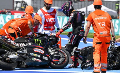 MotoGP pit area with riders and crew surrounding two racing motorcycles on a blue track surface.