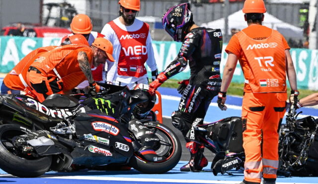 MotoGP pit area with riders and crew surrounding two racing motorcycles on a blue track surface.