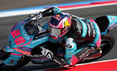 Motorcycle racer in turquoise and pink gear leaning into a sharp turn on a race track with red-and-white curbing behind.