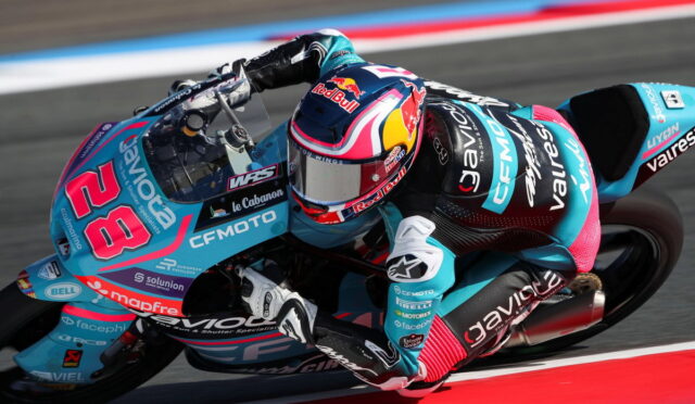 Motorcycle racer in turquoise and pink gear leaning into a sharp turn on a race track with red-and-white curbing behind.
