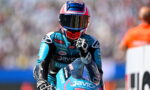 Moto racer in blue racing suit and helmet, fist raised, on a blue bike at a blurred track background.