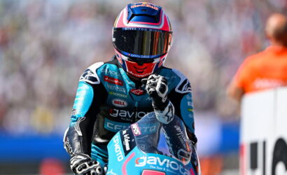 Moto racer in blue racing suit and helmet, fist raised, on a blue bike at a blurred track background.