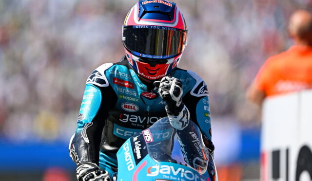 Moto racer in blue racing suit and helmet, fist raised, on a blue bike at a blurred track background.