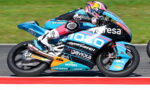 MotoGP rider in blue racing leathers on a sponsor-liveried bike leaning into a turn on a race track.