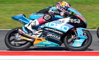 MotoGP rider in blue racing leathers on a sponsor-liveried bike leaning into a turn on a race track.
