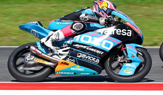 MotoGP rider in blue racing leathers on a sponsor-liveried bike leaning into a turn on a race track.