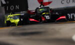 Black and red Formula race car on track with a bright yellow car in the background, emphasizing a high-speed racing moment on a street circuit.