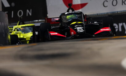 Black and red Formula race car on track with a bright yellow car in the background, emphasizing a high-speed racing moment on a street circuit.