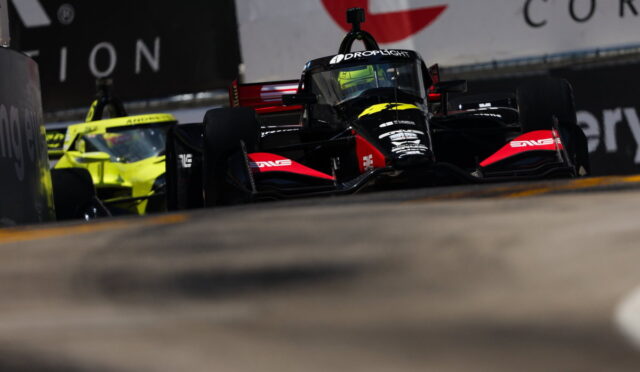 Black and red Formula race car on track with a bright yellow car in the background, emphasizing a high-speed racing moment on a street circuit.