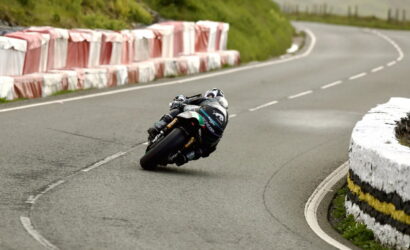 Motorcycle racer leaning sharply into a left-hand bend on a paved road, with red-and-white crash barriers to the left and a white stone barrier to the right.