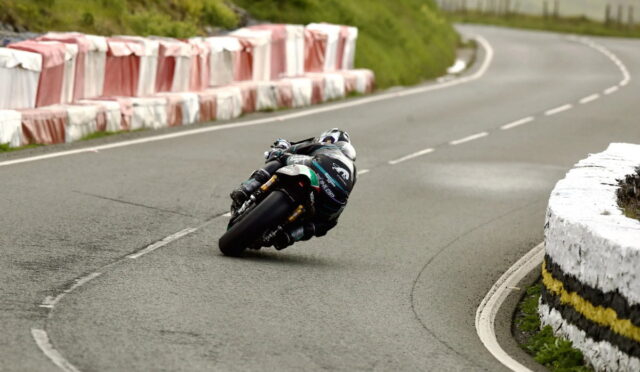 Motorcycle racer leaning sharply into a left-hand bend on a paved road, with red-and-white crash barriers to the left and a white stone barrier to the right.
