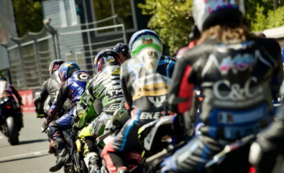 Racers on sport motorcycles lean into a turn from behind, wearing colorful racing suits and helmets on a race track