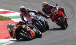 Group of motorcycle racers lean into a sharp turn on a race track, close behind each other on colorful bikes with visible numbers like 38 and 10.