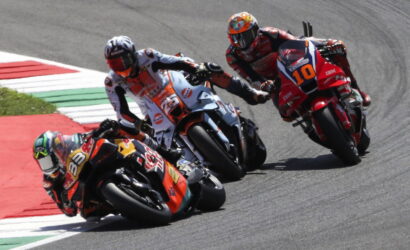 Group of motorcycle racers lean into a sharp turn on a race track, close behind each other on colorful bikes with visible numbers like 38 and 10.