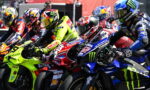 Group of motorcycle racers leaning into a tight start on a race track, colorful bikes and helmets close together.