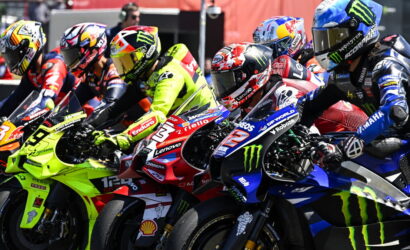Group of motorcycle racers leaning into a tight start on a race track, colorful bikes and helmets close together.