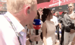 Close-up of a reporter holding a blue-red 'Sports' microphone, interviewing a woman in a beige dress at Monaco Circuit.