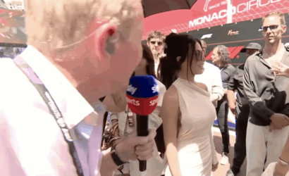Close-up of a reporter holding a blue-red 'Sports' microphone, interviewing a woman in a beige dress at Monaco Circuit.