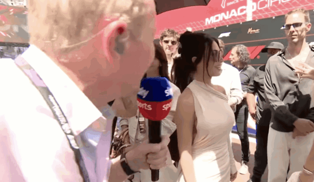 Close-up of a reporter holding a blue-red 'Sports' microphone, interviewing a woman in a beige dress at Monaco Circuit.