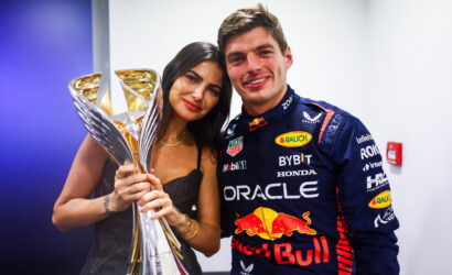 Race car driver in a blue Red Bull Honda suit posing with a tall golden trophy beside a smiling woman.