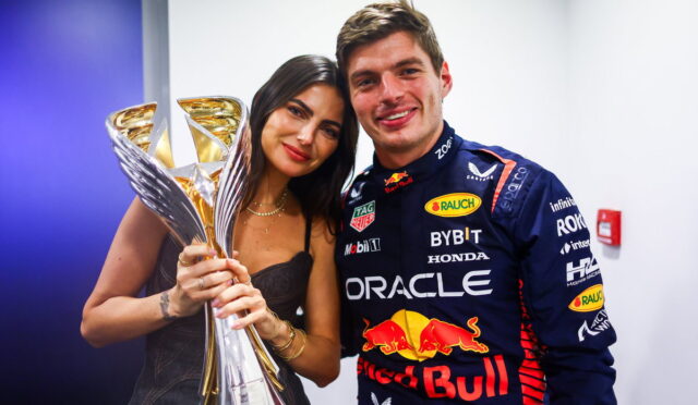 Race car driver in a blue Red Bull Honda suit posing with a tall golden trophy beside a smiling woman.