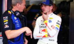 Two Formula 1 crew members chat in a garage, one in a white Red Bull racing suit and cap, the other in a blue team shirt with headphones nearby.